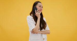 Woman in a white blazer talking on a cell phone against a solid yellow background with a thoughtful expression