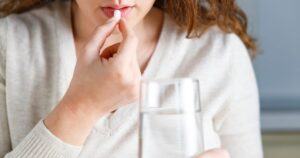 Woman placing a white pill in her mouth with a glass of water nearby