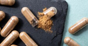 Brown powder spilled from clear capsule on a dark slate surface surrounded by other filled capsules herbal supplement