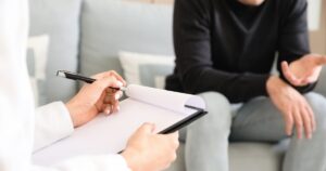 Person writes on a clipboard while a client sits on a sofa and gestures during a discussion