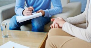 Therapist taking notes on a clipboard while a client sits with hands clasped in a counseling session at home