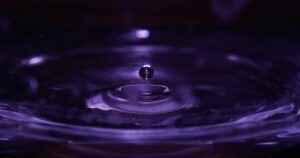 Close up of a single water droplet hovering above a rippling purple tinted pool surface just before impact