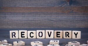 Wooden letter blocks spell RECOVERY on a dark rustic table with a few tiles scattered nearby