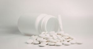 White prescription bottle tipped over with round white pills spilling onto a white surface and lid nearby