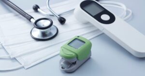 Green digital pulse oximeter with a stethoscope and disposable masks on a light blue surface suggesting medical monitoring gear nearby