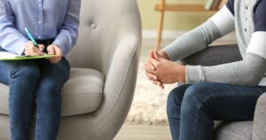Therapist takes notes on a clipboard while client sits with hands clasped in a counseling room