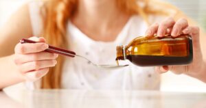 Person pours brown syrup from a bottle into a spoon held over a white surface