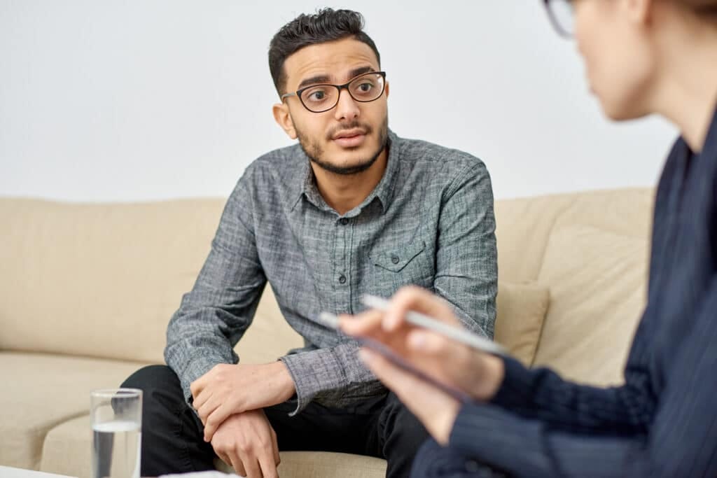 Man with glasses sits on a beige sofa talking to a counselor who is taking notes or writing on a tabletclipboard in the foreground