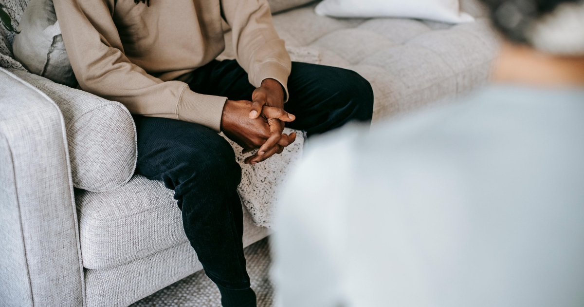 Two people sit on a light gray sofa foreground person blurred while the other rests their hands together in their lap wearing a beige top and dark jeans