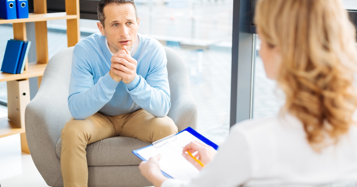 Man in a blue sweater sits in a lounge chair hands folded speaking with a counselor in a bright office with a clipboard nearby