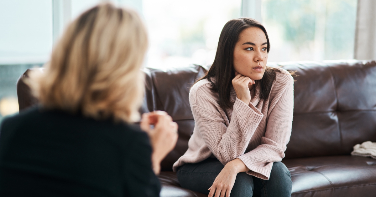 Woman in a pink sweater sits on a brown leather sofa listening thoughtfully to someone speaking nearby in a living room setting