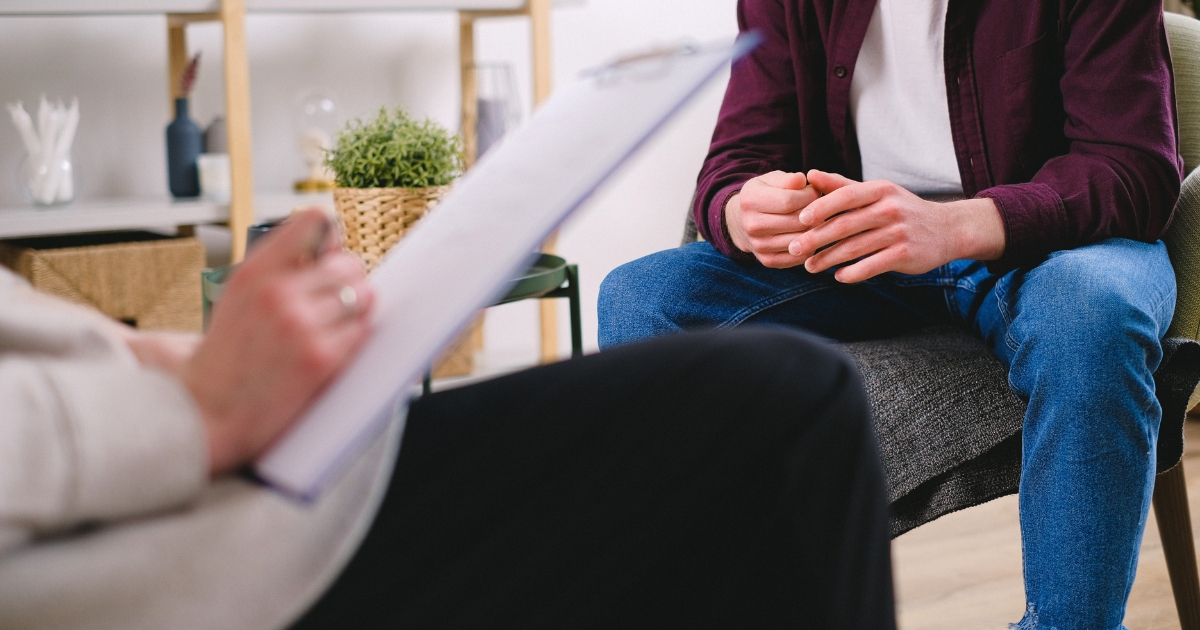 Counselor taking notes on a clipboard while a client sits across hands clasped in a therapy room
