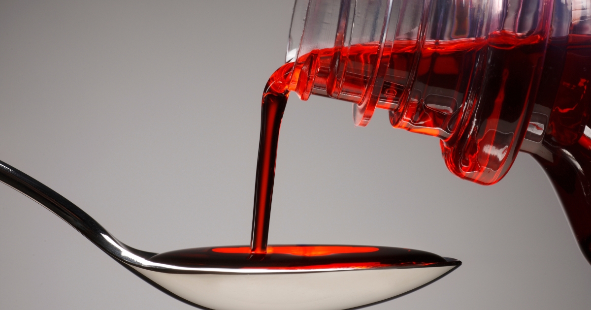 Red syrup pouring from a bottle into a metal spoon with a smooth gray background
