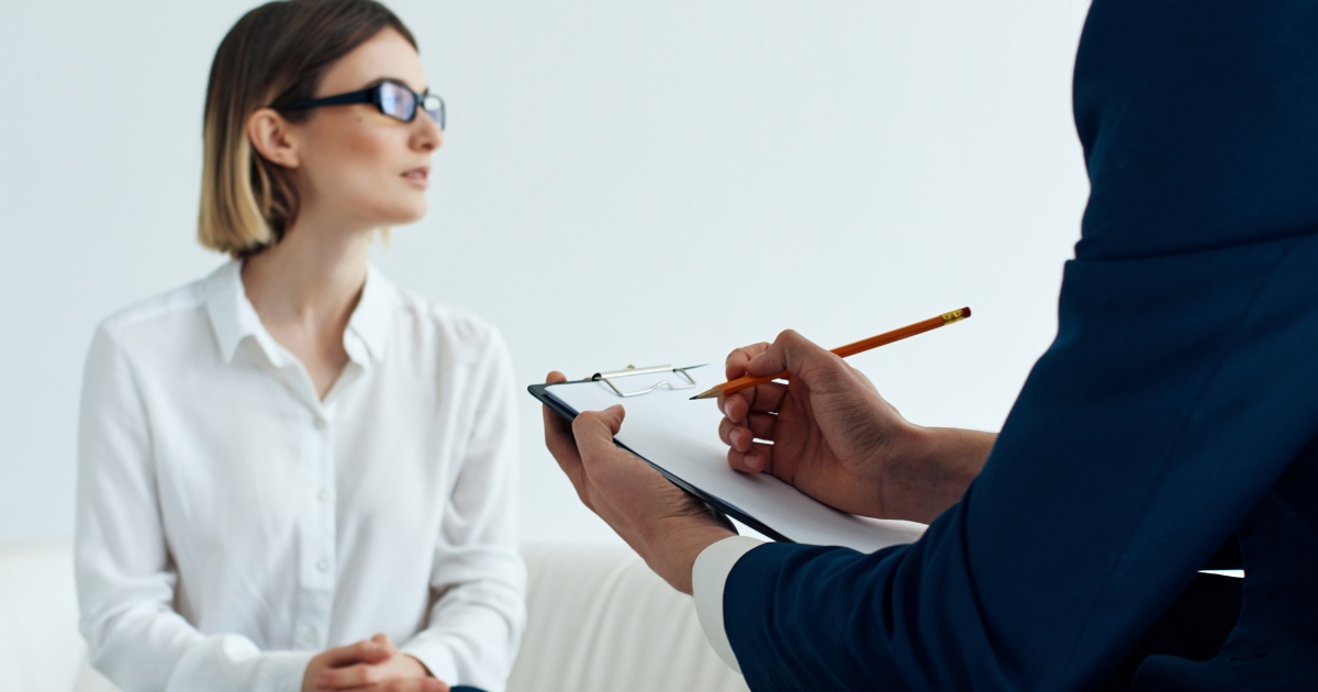 Woman in a white blouse sits on a sofa while a colleague in a dark suit takes notes on a clipboard