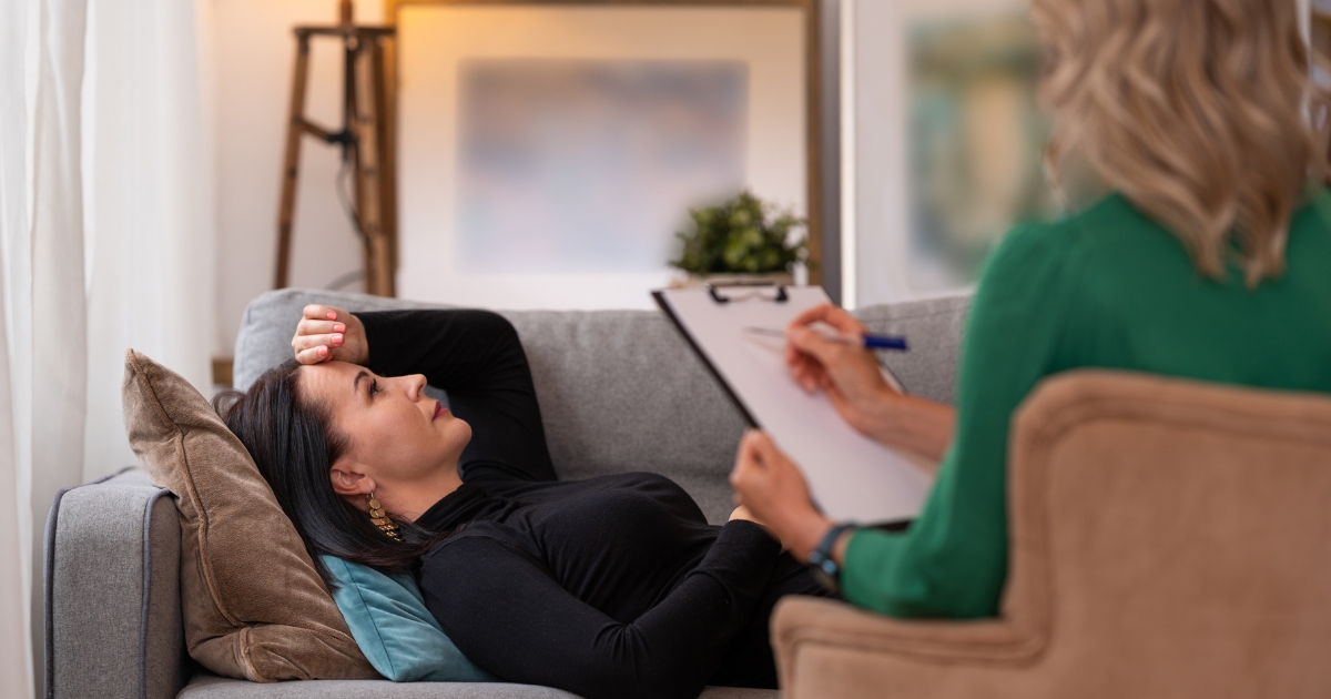 Therapist sits beside a couch taking notes on a clipboard while a woman lies on the sofa talking