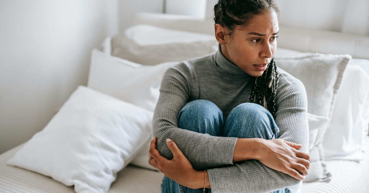 Woman sits on a bed knees drawn in and arms wrapped around them with a worried expression in a bright bedroom setting