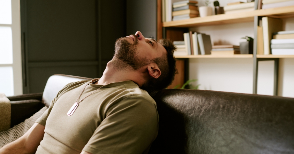 Man leaning back on a dark leather sofa head tilted up wearing a dog tag necklace in a living room with bookshelves behind him