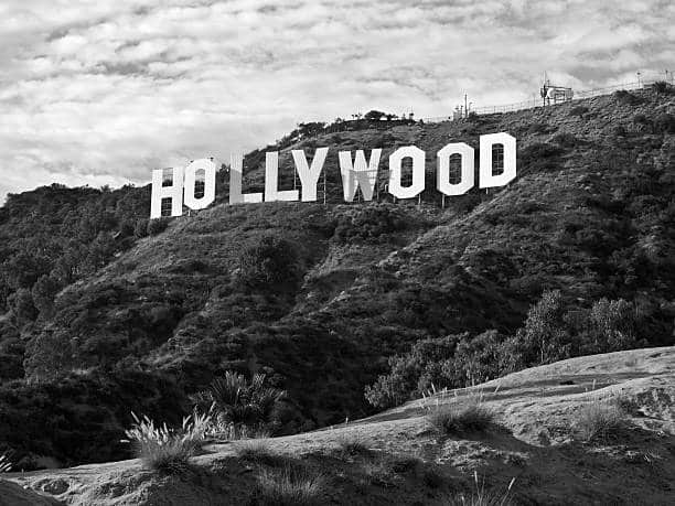 Black and white photo of the Hollywood Sign perched on a hillside with rugged terrain and a cloudy sky