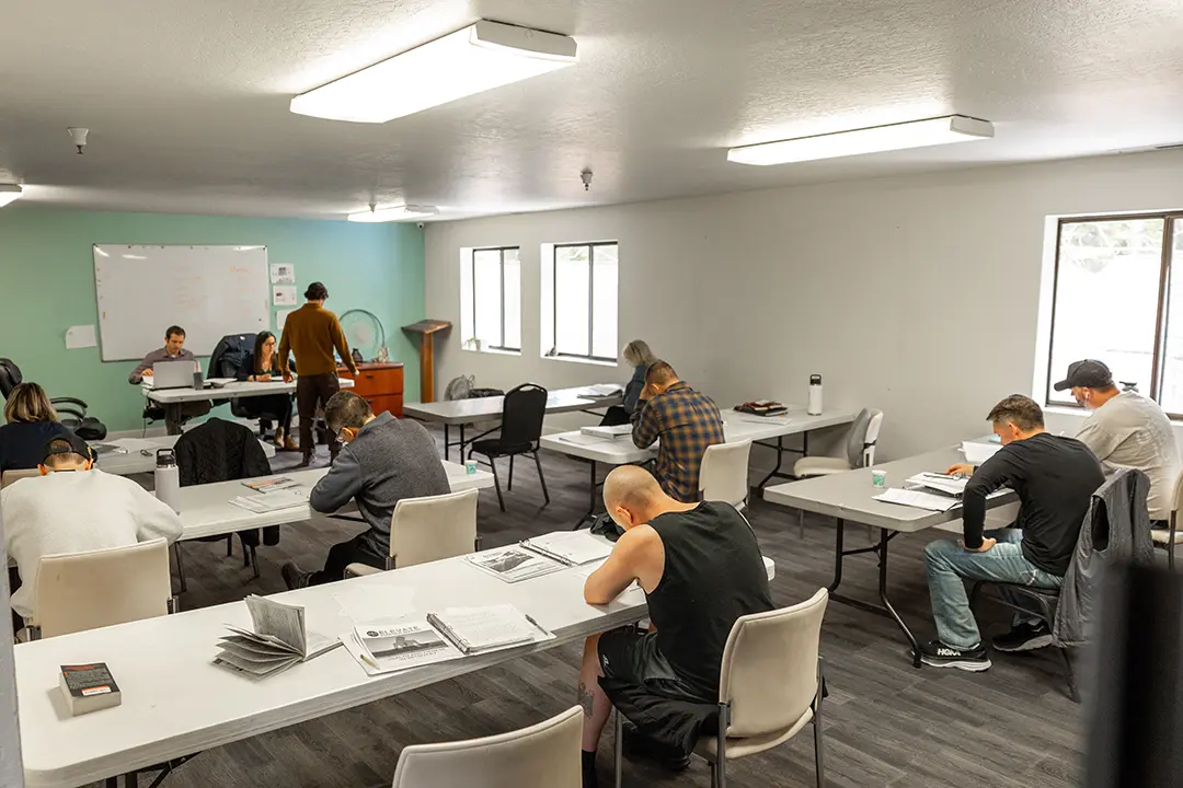 Adult students sit at long tables in a classroom taking notes as an instructor stands near a whiteboard at the front