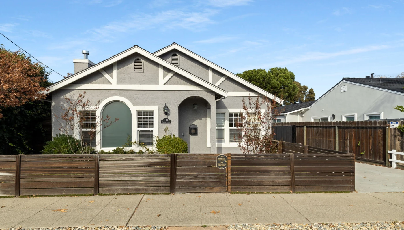 Front view of a single story gray house with white trim a wooden privacy fence and a small front garden under a blue sky