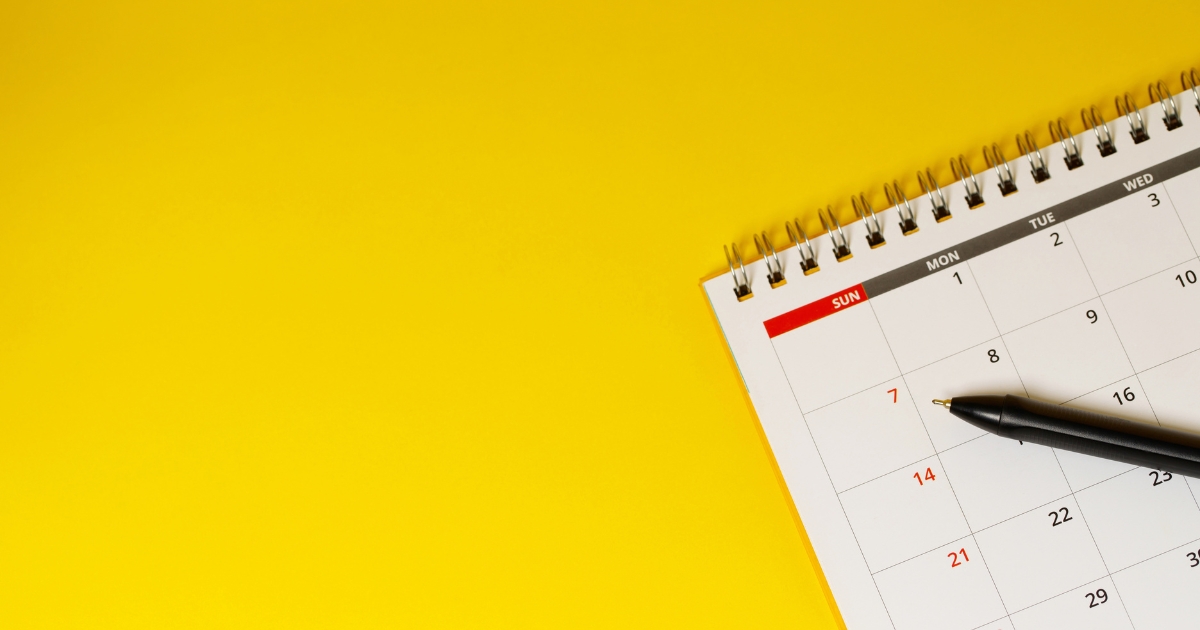Close up of a spiral calendar on a bright yellow background with a black pen resting on it ready to mark dates