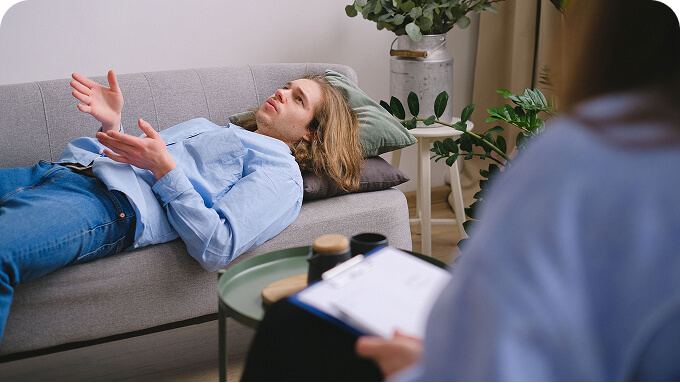 Man with long blond hair lying on a gray sofa speaking during a therapy session with a clinician taking notes nearby