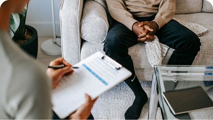 Therapist seated left writing on a clipboard while a client sits crosslegged on a sofa hands folded in a living room setting