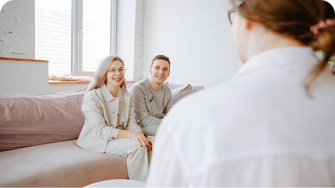 Two people sit on a pale pink couch in a bright office like room smiling toward the person in front of them A third person is visible from behind