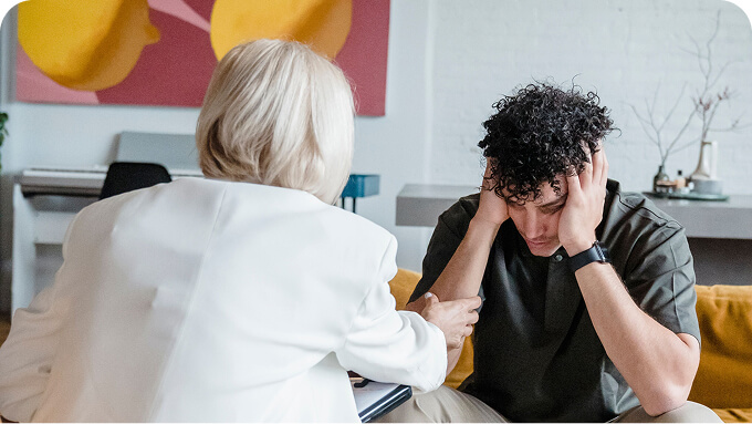 Counselor in a white blazer sits beside a distressed young man on a couch offering support