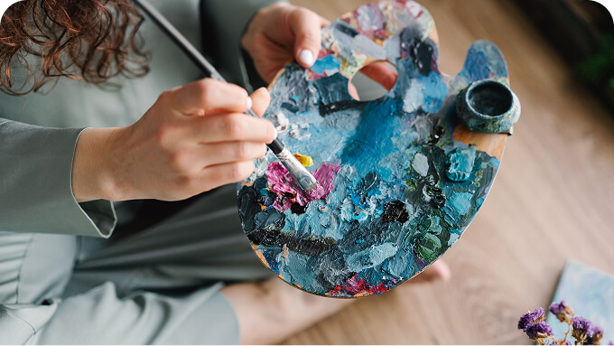 Close up of a person holding a paint palette covered in blue and other colors painting with a brush