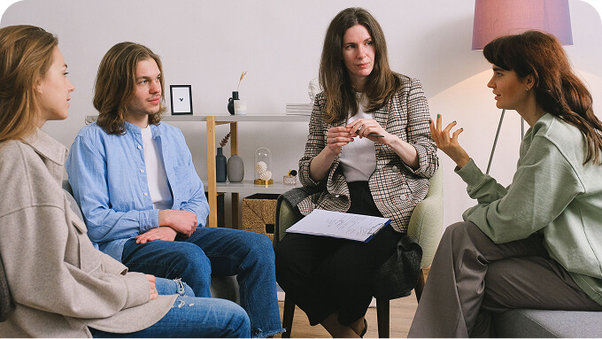Group therapy session with four adults seated in a circle facilitator woman speaks while others listen one has a notebook and pen nearby