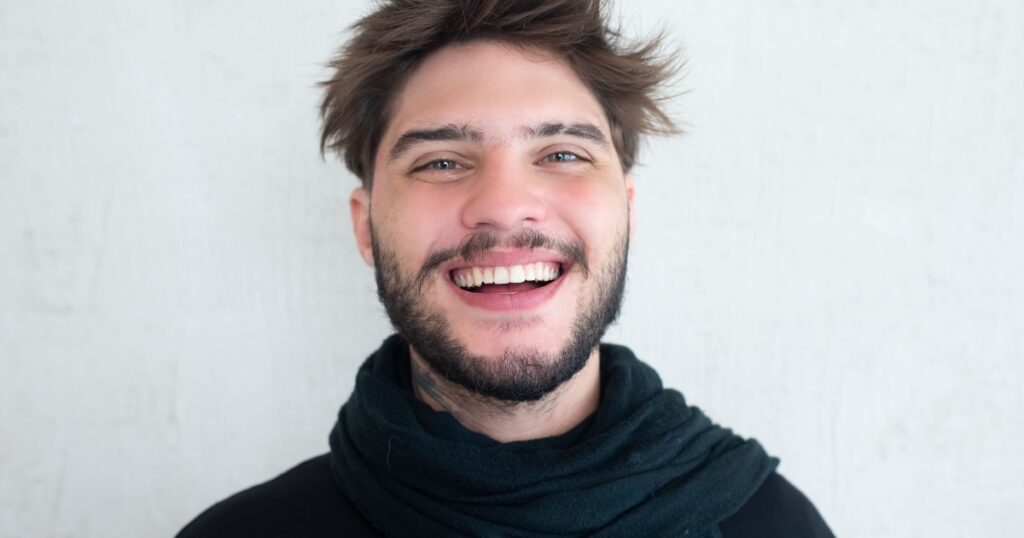 Man with a beard and short brown hair smiling at the camera wearing a dark scarf against a light gray wall