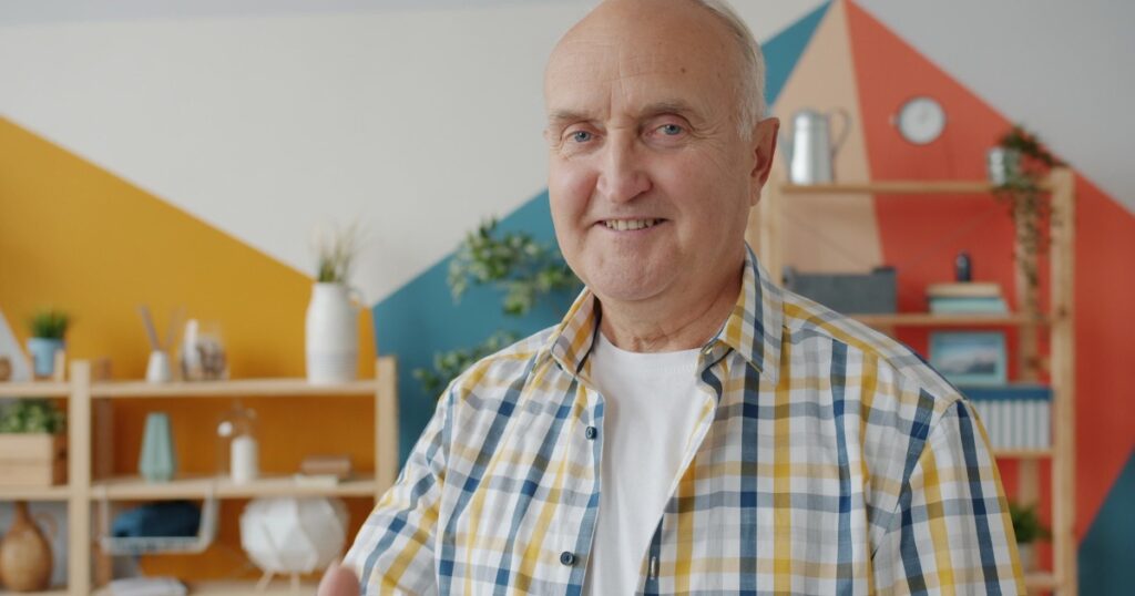 Smiling older man in a plaid yellow blue shirt standing in a bright room with geometric orange and blue wall art and shelves