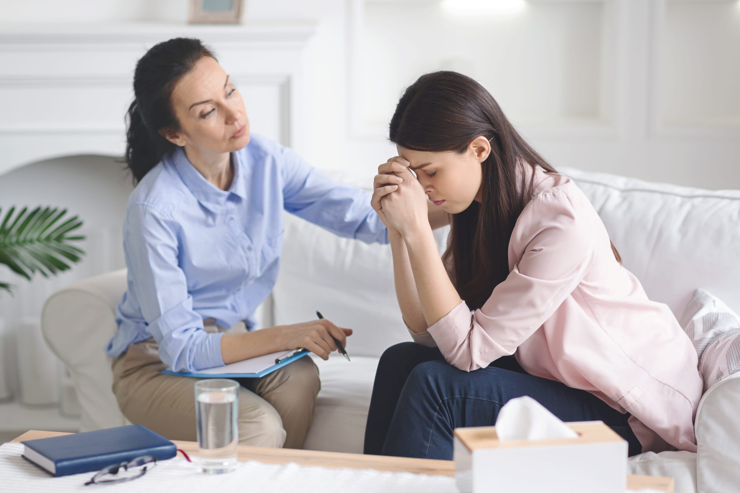 Therapist writes on a clipboard while a worried woman sits nearby holding her head in her hands in a living room setting