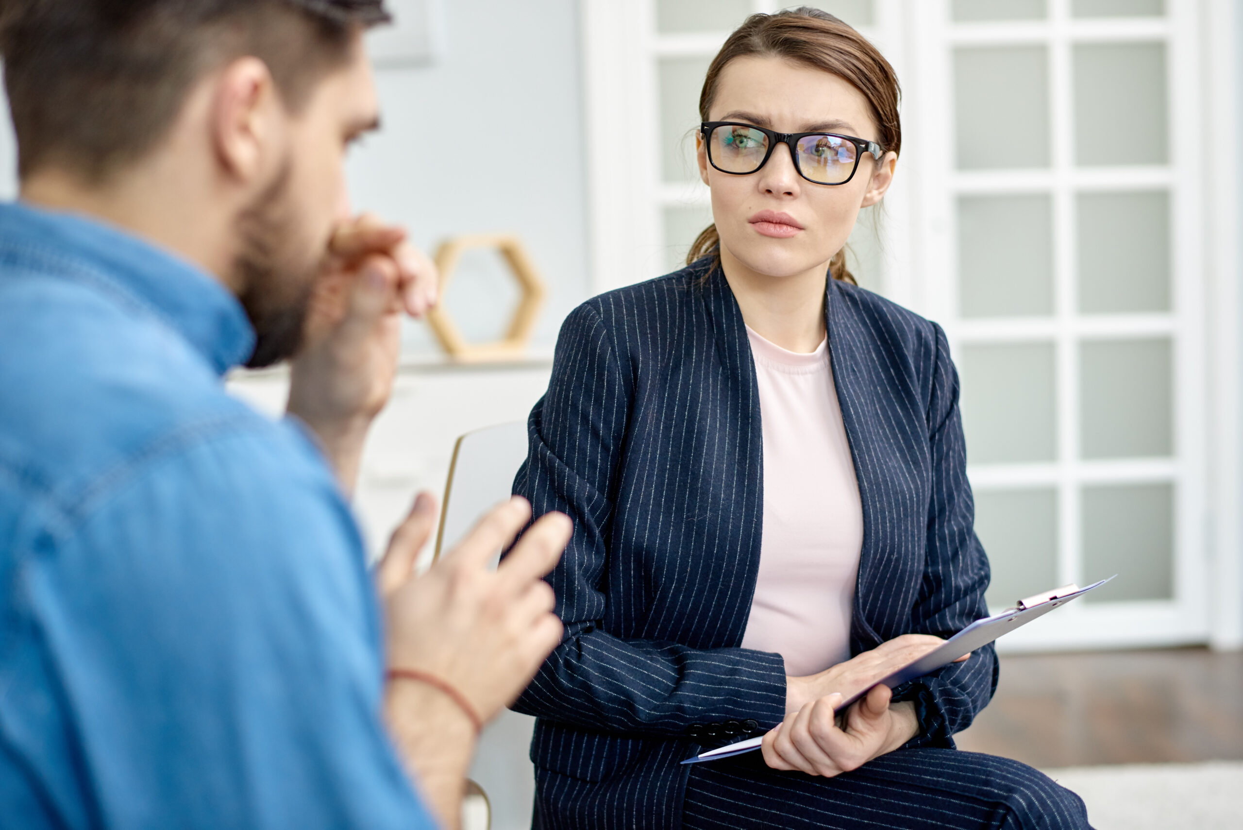 Professional woman in a pinstripe suit and pink blouse holds a clipboard while talking to a man in the background on a meeting