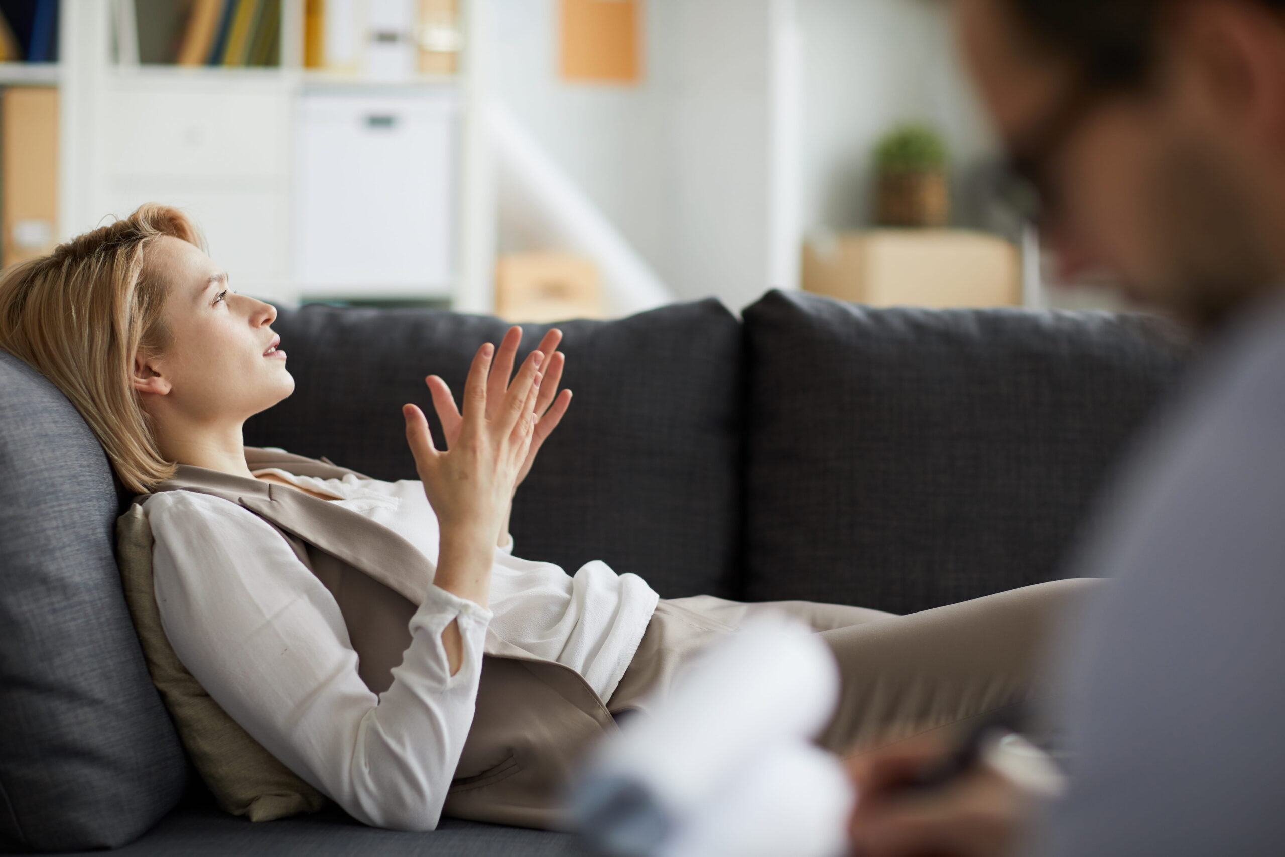 Woman resting on a gray sofa gesturing with raised hands while talking to an unseen person in a living room
