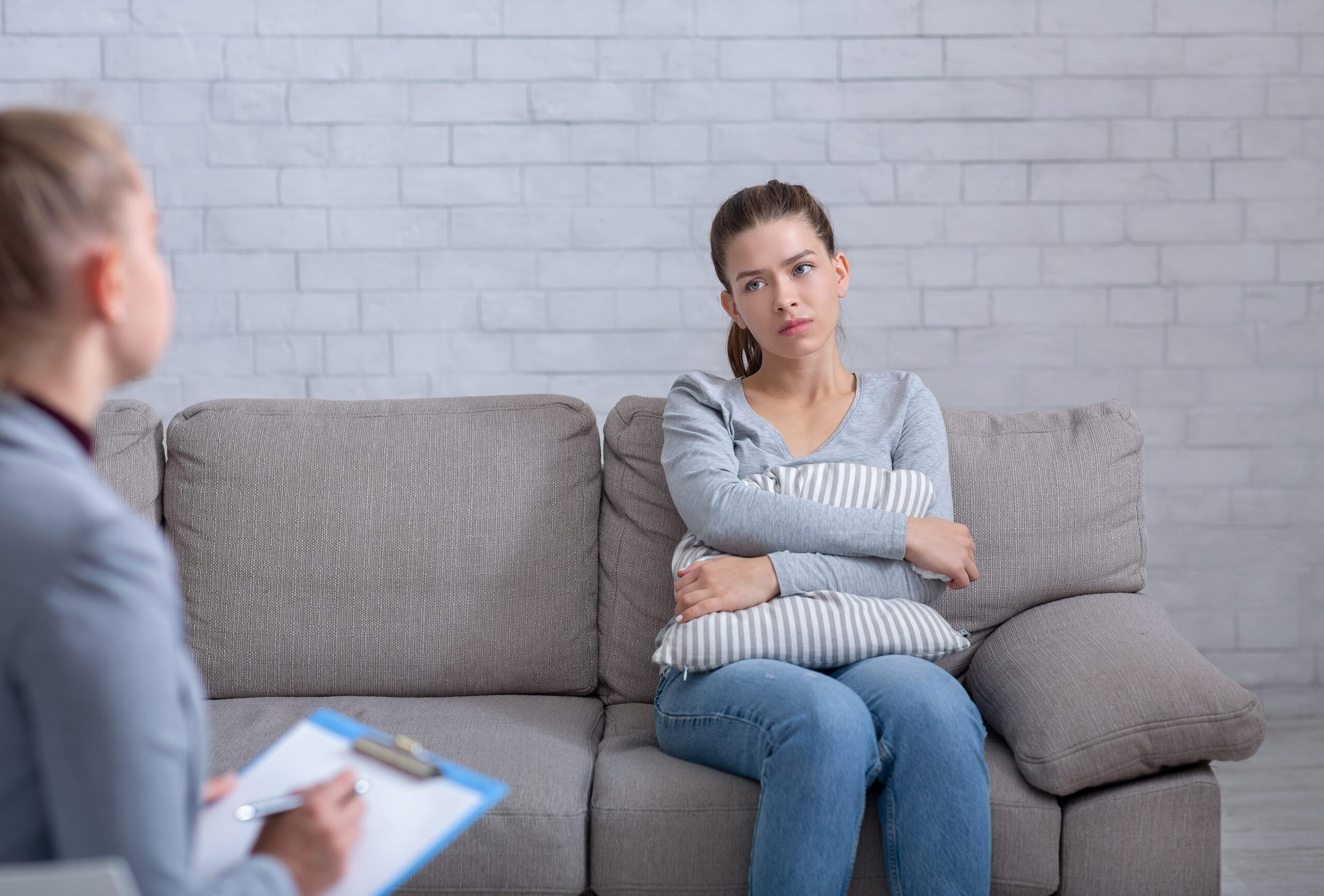 Woman sitting on a gray sofa with folded arms listening during a therapy session therapist with a clipboard is in the foreground blurred